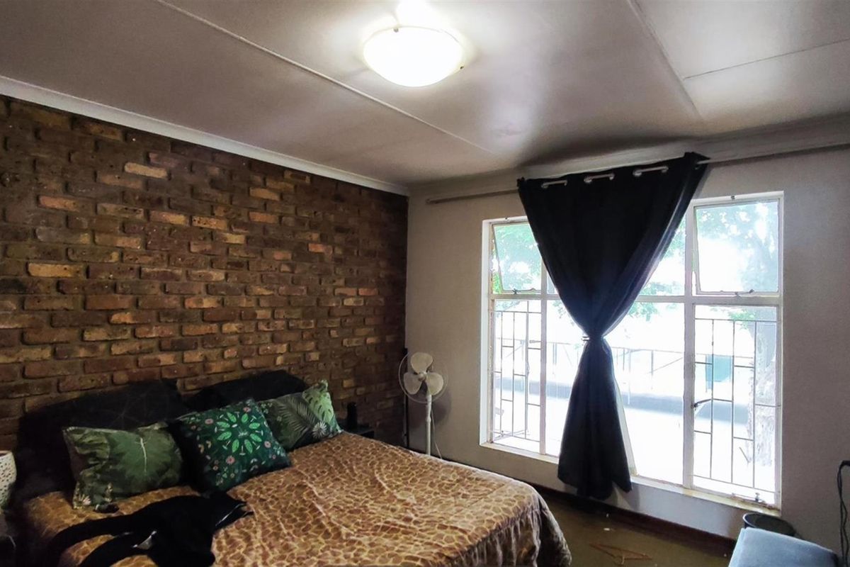 A bedroom featuring a rustic exposed brick feature wall, large window with security bars, and a ceiling-mounted light fixture.