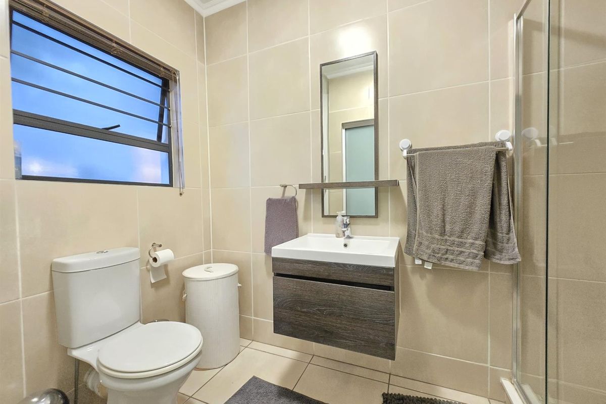 A modern bathroom featuring floor-to-ceiling beige tiling, a floating vanity with storage, a glass-enclosed shower, and a window with security bars.