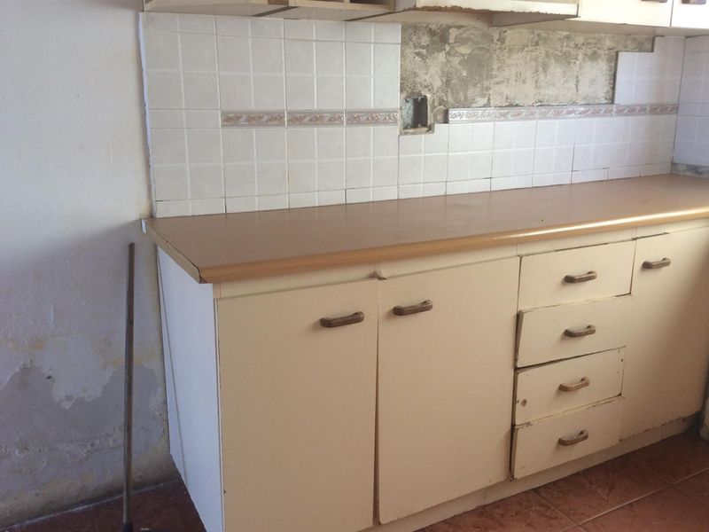 A dated kitchen area featuring original cabinetry, laminate countertops, and tiled backsplash, currently requiring significant renovation and repair to the wall finishes.