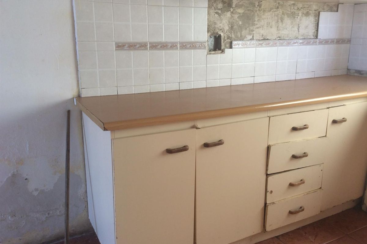 A dated kitchen area featuring original cabinetry, laminate countertops, and tiled backsplash, currently requiring significant renovation and repair to the wall finishes.