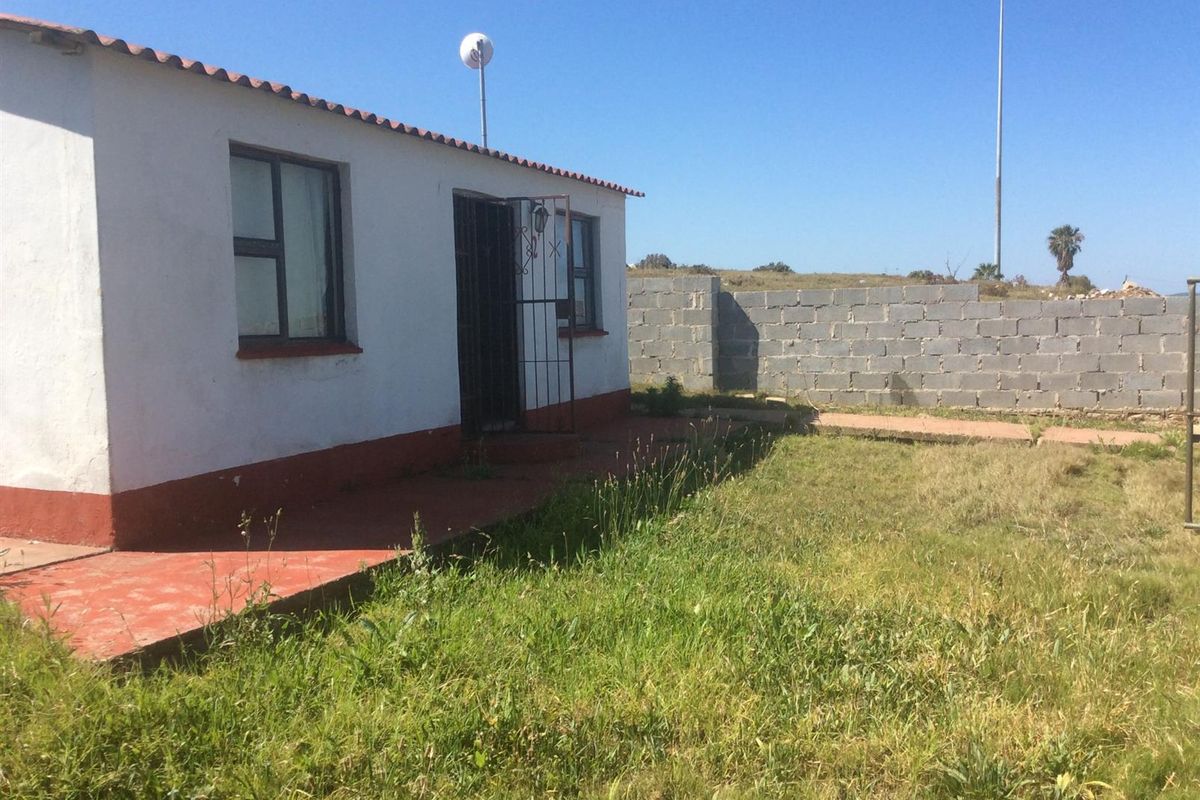 Rear view of the property featuring a small concrete patio, security gate, and an unfinished concrete block boundary wall with an overgrown lawn.