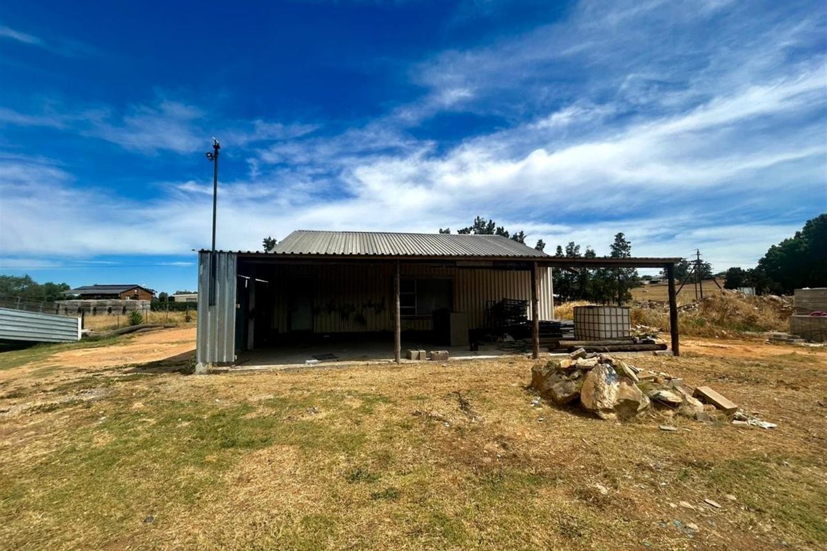 A basic metal-clad structure on a vacant residential plot, featuring a covered patio area and open surrounding land.