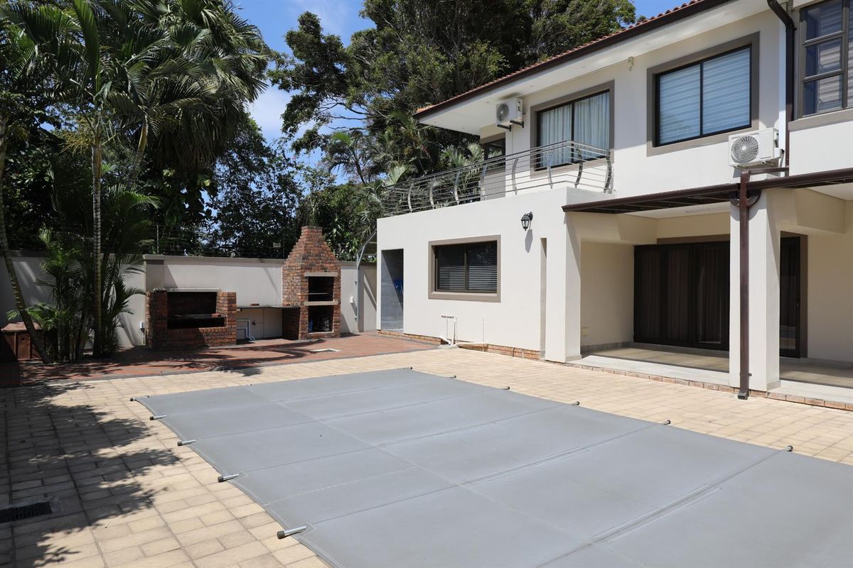 Rear exterior view featuring a paved pool area with a covered swimming pool, built-in brick braai, and a balcony overlooking the garden.