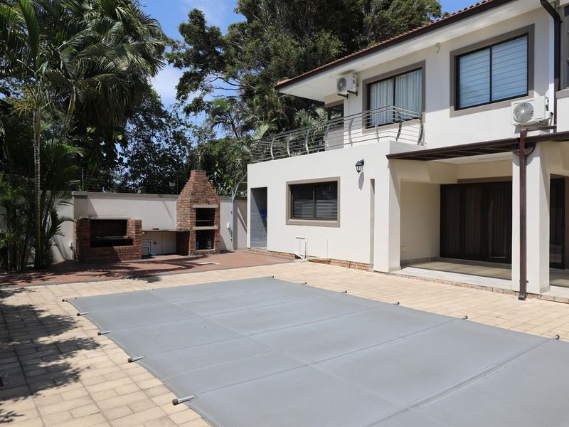 Rear exterior view featuring a paved pool area with a covered swimming pool, built-in brick braai, and a balcony overlooking the garden.