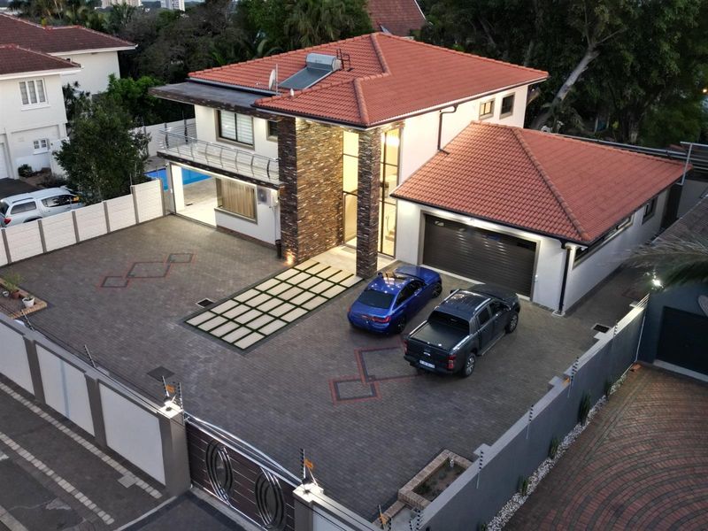 Aerial view of a modern double-story house featuring a tiled roof, stone-clad facade, paved driveway, double garage, and perimeter security fencing.