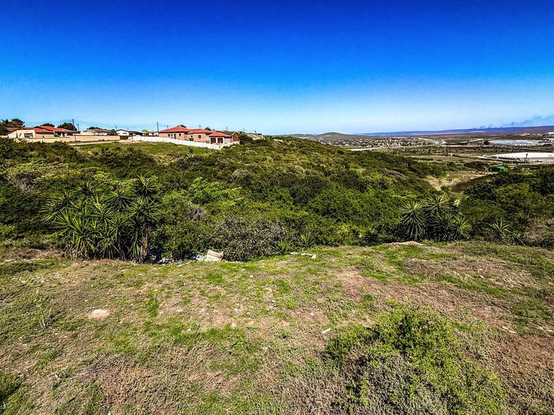A wide-angle view of a vacant residential plot featuring natural vegetation, rolling terrain, and distant neighborhood views under a clear blue sky.