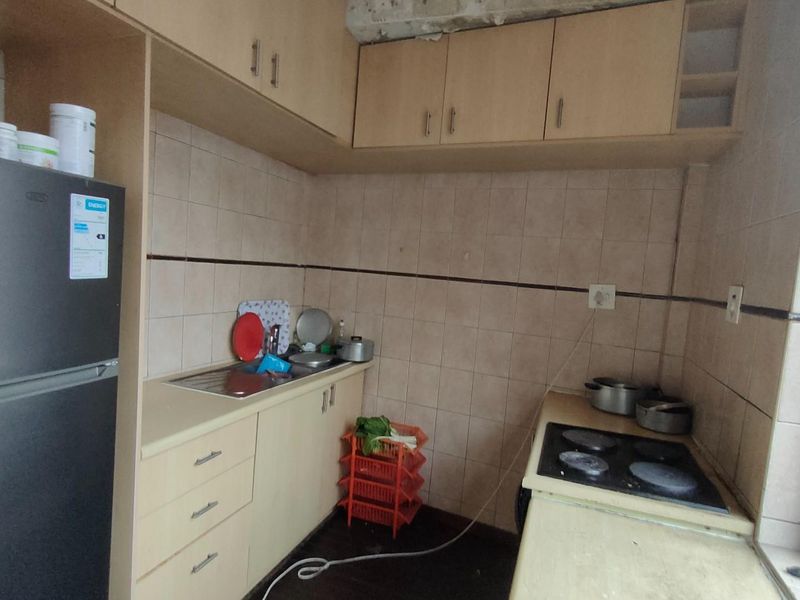 Compact kitchen featuring light wood-tone cabinetry, integrated electric hob, stainless steel sink, and tiled splashbacks in need of cosmetic updates.