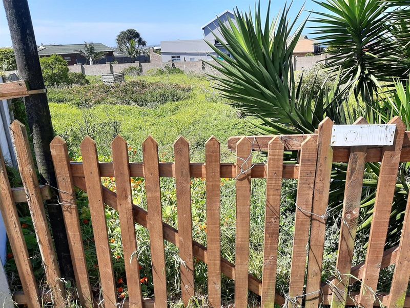 A view of a vacant residential plot featuring an overgrown garden, enclosed by a weathered wooden picket gate and perimeter walling.