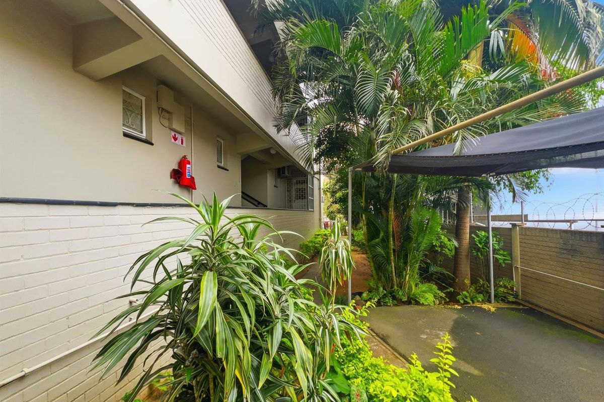 Exterior view of the apartment building featuring a paved walkway, lush garden landscaping, and a shaded outdoor area.