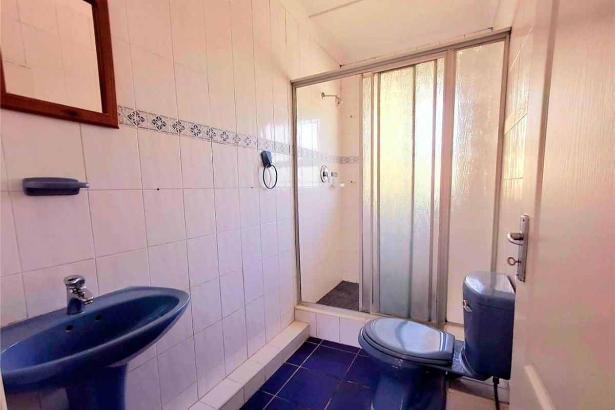 A dated bathroom featuring a blue pedestal basin, matching toilet, and a glass-enclosed shower with tiled walls and flooring.