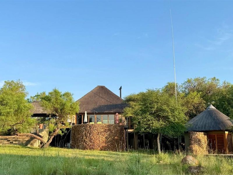 Rear view of a bushveld-style property featuring traditional thatched roofing, natural stone cladding, and an elevated wooden deck set within a natural landscape.