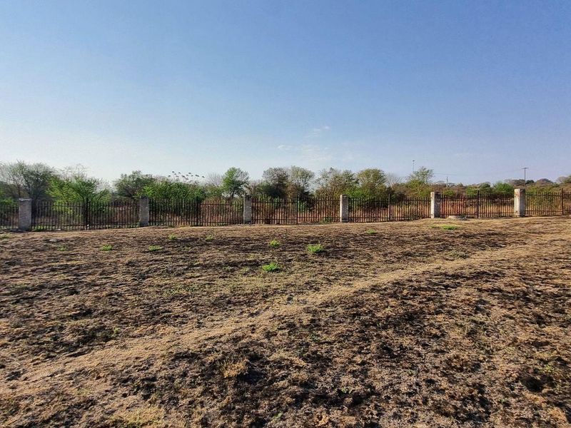 A view of a vacant residential plot featuring dry ground and a perimeter metal fence with concrete posts.