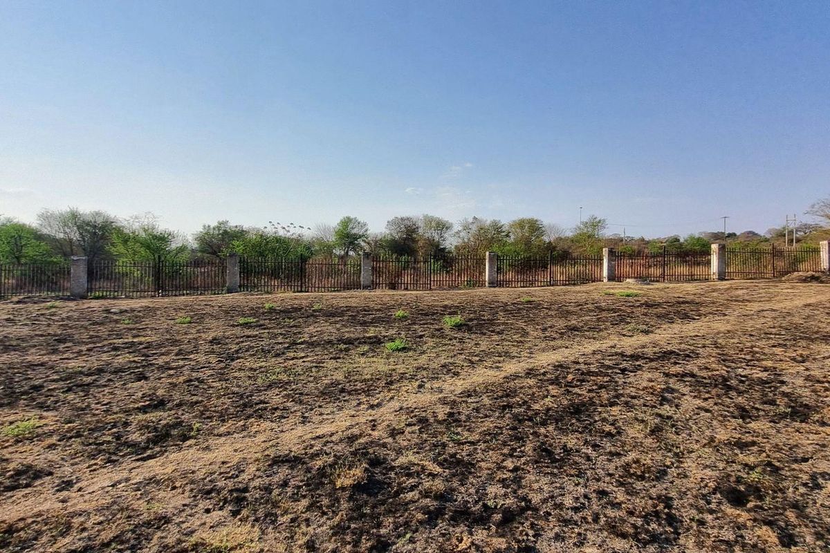 A view of a vacant residential plot featuring dry ground and a perimeter metal fence with concrete posts.