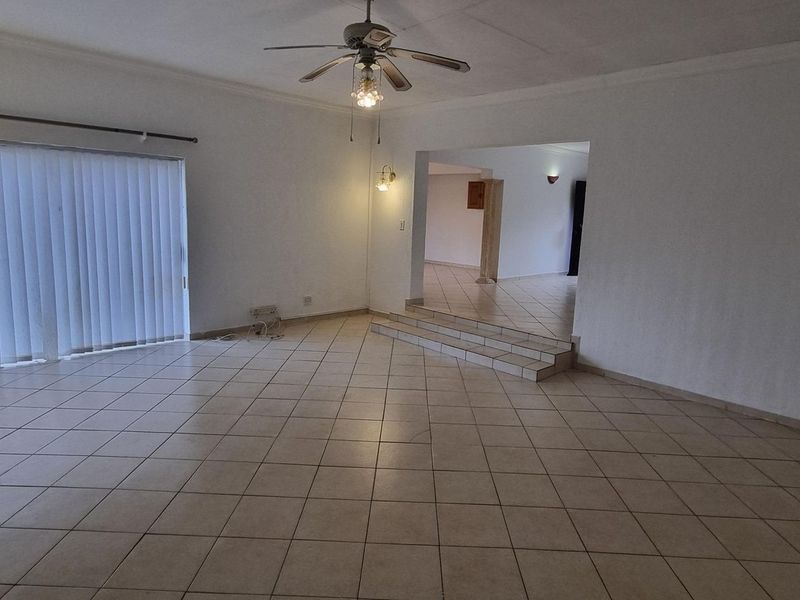 Spacious, open-plan living area featuring neutral tiled flooring, a ceiling fan, and a raised platform leading to an adjacent room.