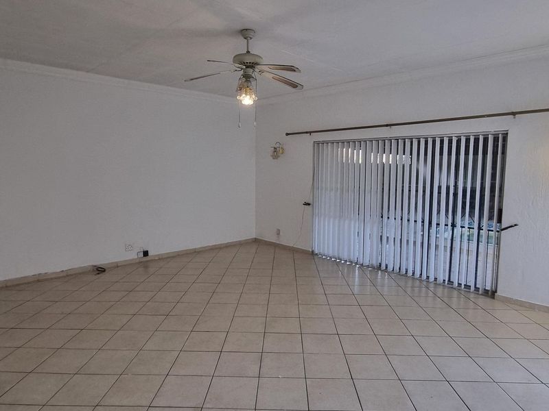 Spacious, empty living room featuring neutral tiled flooring, a ceiling fan, and large sliding glass doors with vertical blinds leading to the exterior.
