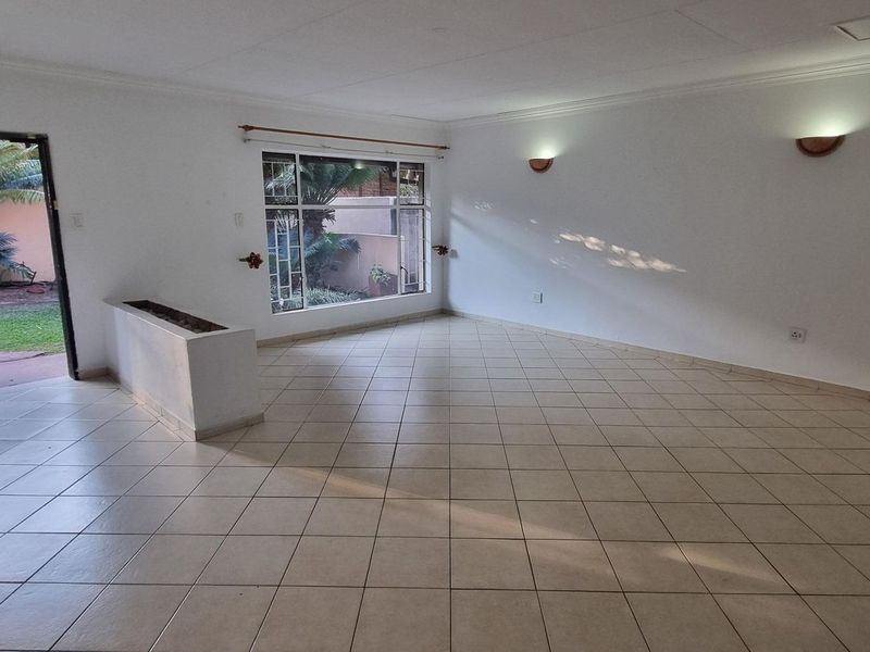 A spacious, empty living area featuring neutral tiled flooring, wall-mounted lighting, and a large window with direct access to the garden.