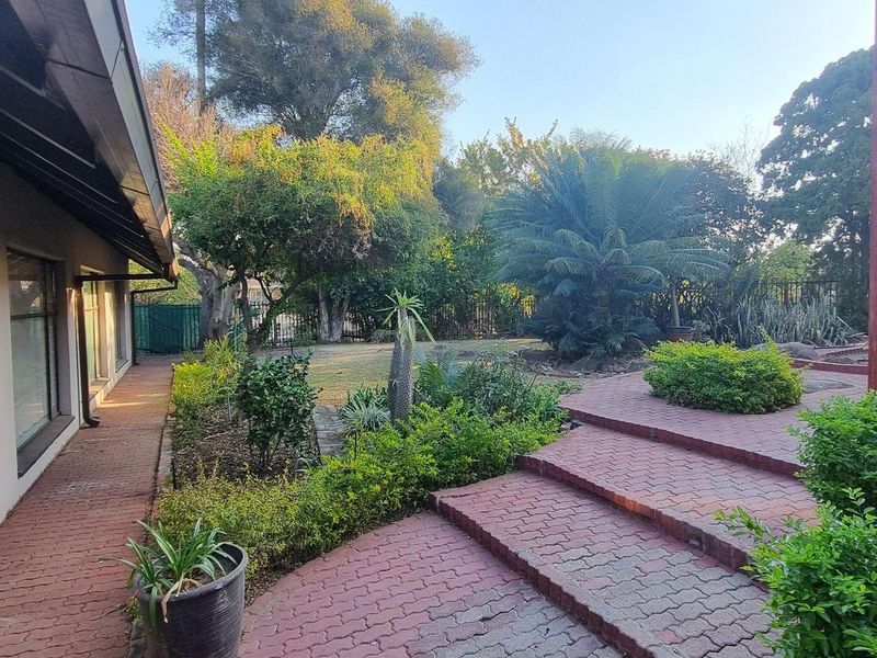 A spacious rear garden featuring a brick-paved patio area with steps, established landscaping, and a covered walkway along the side of the house.