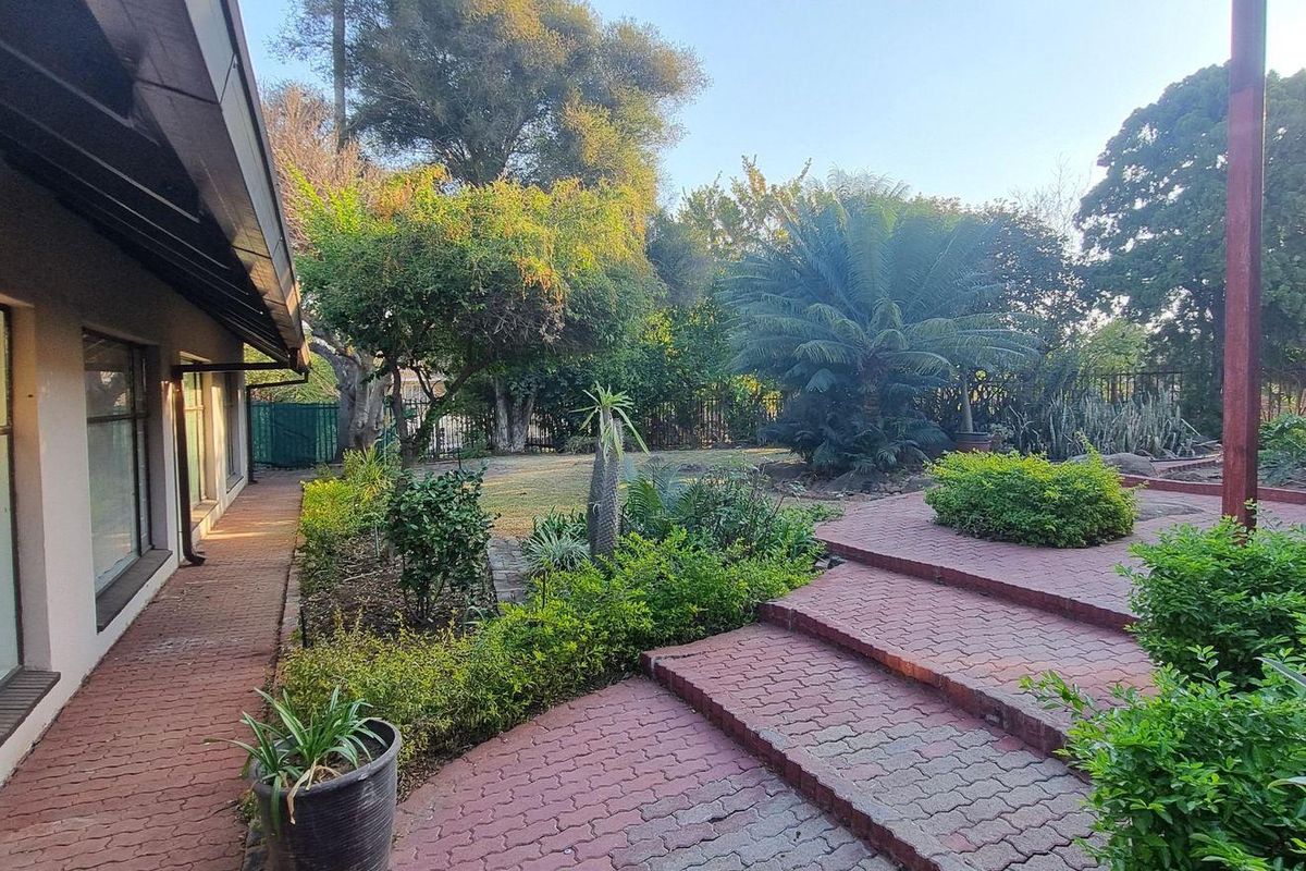 A spacious rear garden featuring a brick-paved patio area with steps, established landscaping, and a covered walkway along the side of the house.