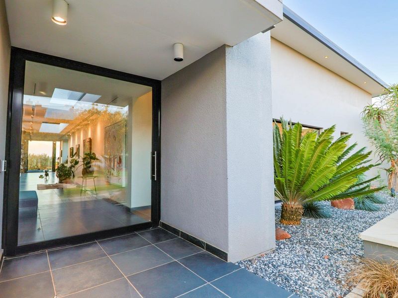 Modern entrance featuring a large glass pivot door, recessed ceiling lighting, and tiled porch area with landscaped garden surrounds.
