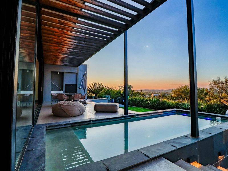 Modern outdoor entertainment area featuring a sleek swimming pool, covered timber pergola, and built-in braai, offering expansive sunset views.