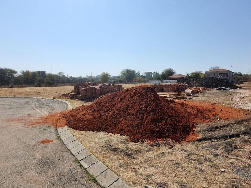 A vacant residential plot featuring a paved driveway, perimeter fencing, and ongoing construction of a brick building structure.