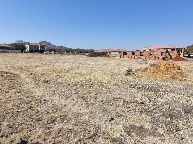 A wide-angle view of a vacant residential plot featuring dry grass, uneven terrain, and surrounding neighborhood development.