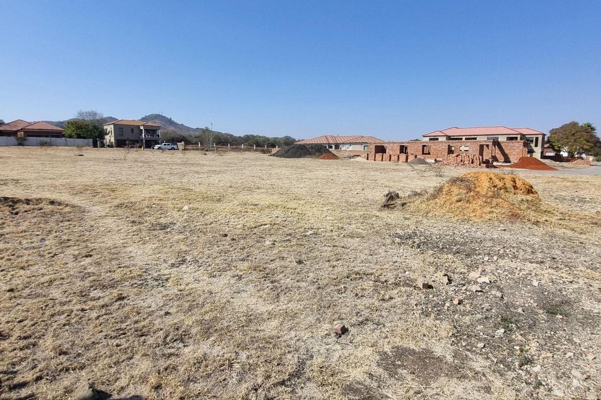 A wide-angle view of a vacant residential plot featuring dry grass, uneven terrain, and surrounding neighborhood development.