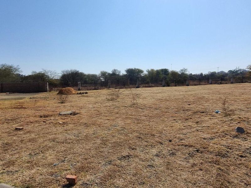 A wide, open residential plot featuring dry grass, perimeter fencing, and a partial view of an adjacent building structure.