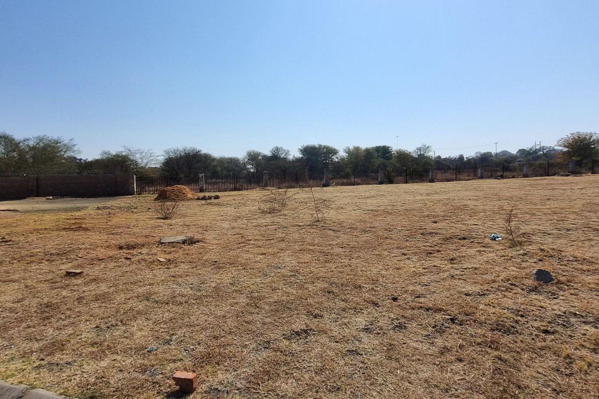 A wide, open residential plot featuring dry grass, perimeter fencing, and a partial view of an adjacent building structure.