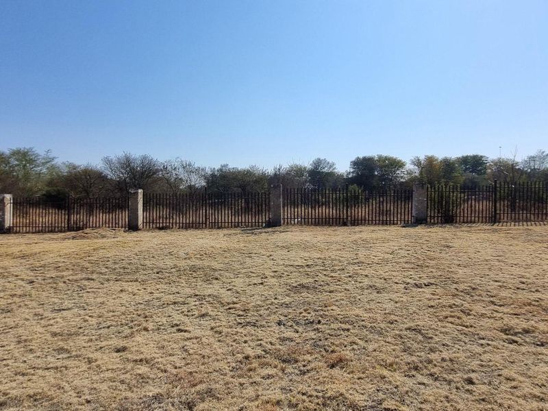 A vacant residential plot featuring dry grass and a perimeter metal fence with masonry posts.
