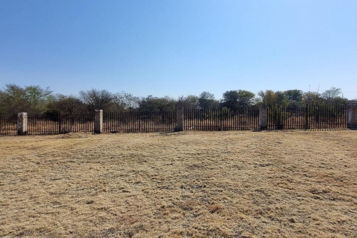 A vacant residential plot featuring dry grass and a perimeter metal fence with masonry posts.