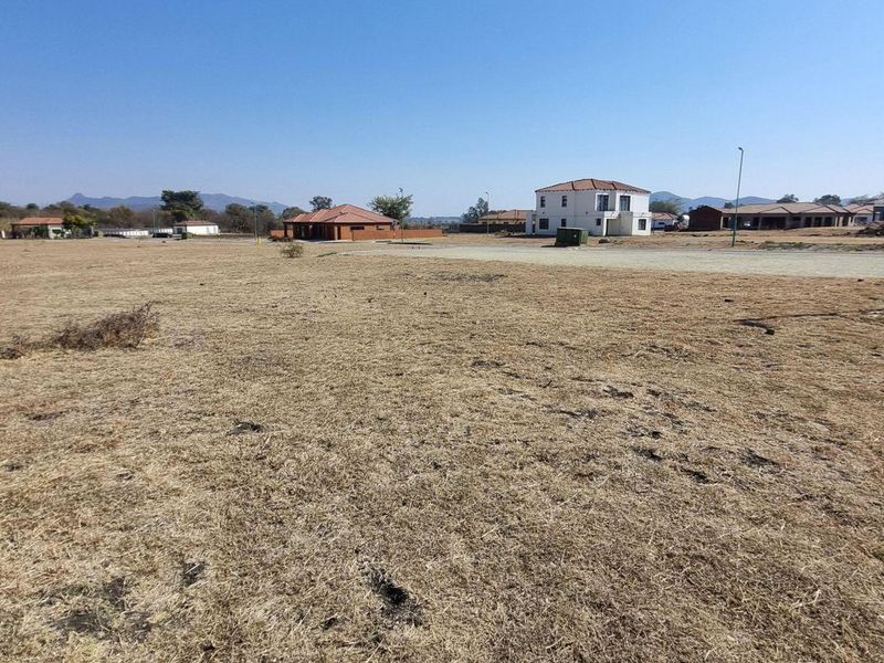 A wide-angle view of a vacant residential plot in a developed estate, featuring dry grass and direct access to a paved road.