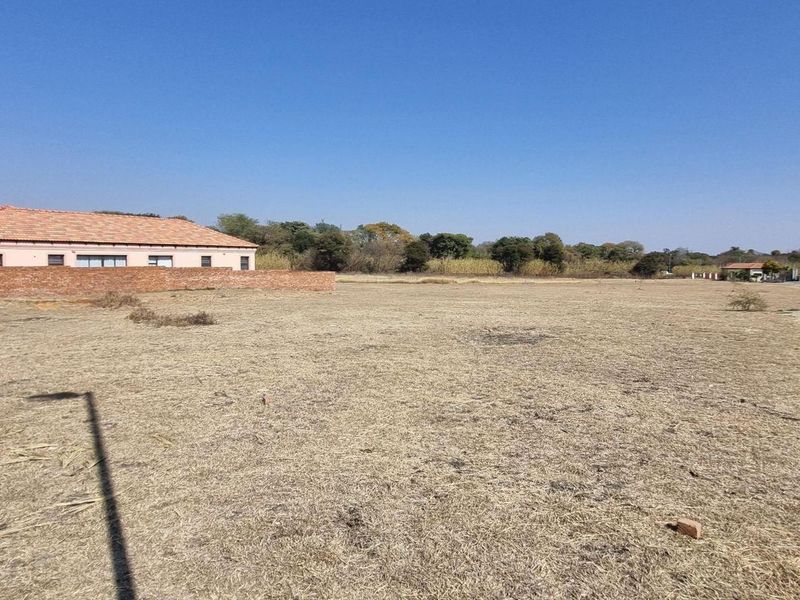 A wide-angle view of a vacant residential plot featuring dry grass, a boundary wall, and an external water tap.