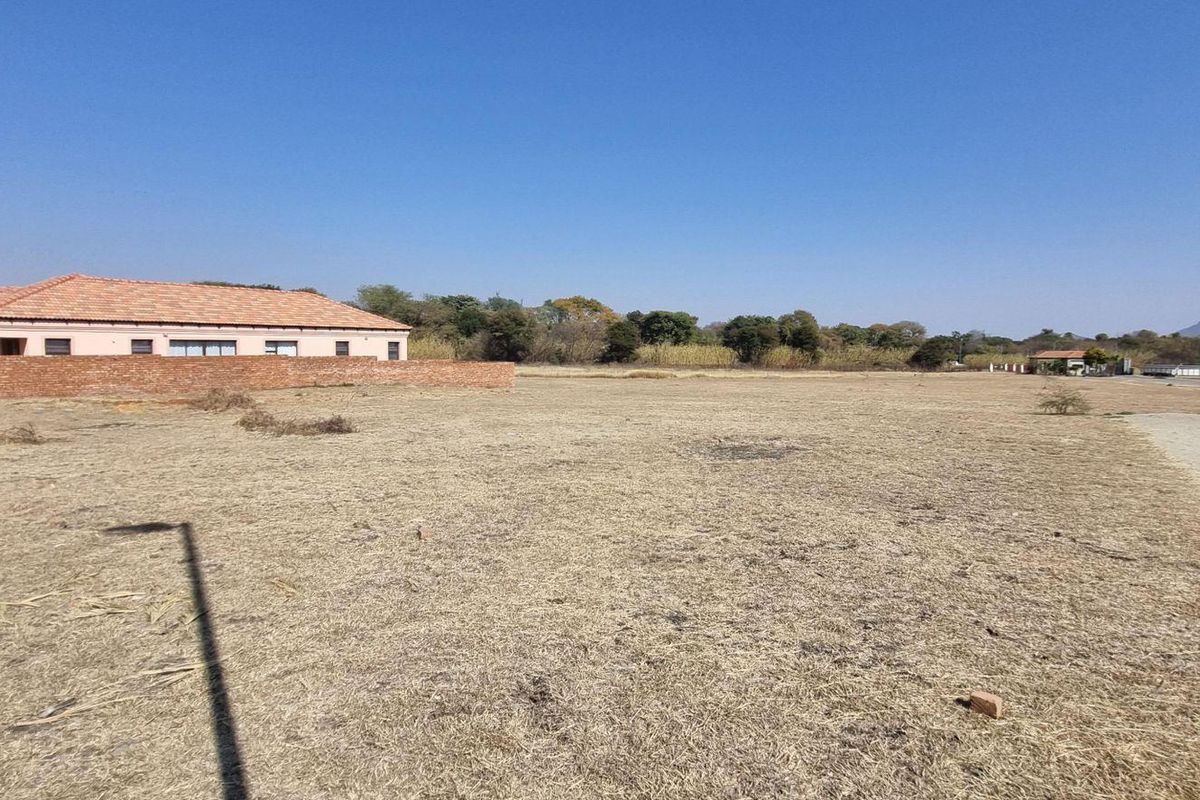 A wide-angle view of a vacant residential plot featuring dry grass, a boundary wall, and an external water tap.