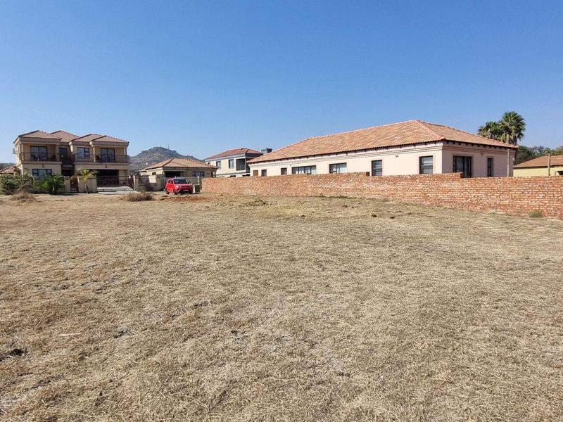 A vacant residential plot featuring dry grass and a brick boundary wall, situated in a developed neighborhood with surrounding residential properties.