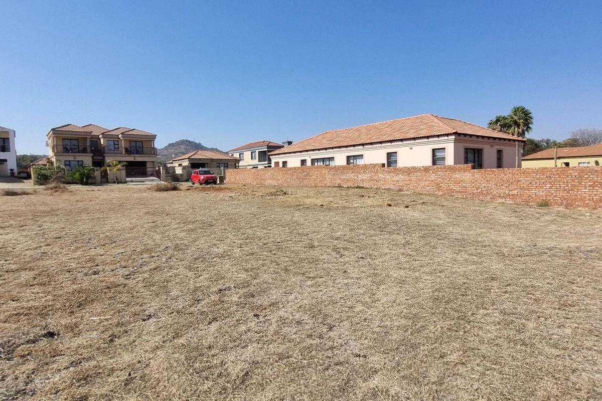 A vacant residential plot featuring dry grass and a brick boundary wall, situated in a developed neighborhood with surrounding residential properties.