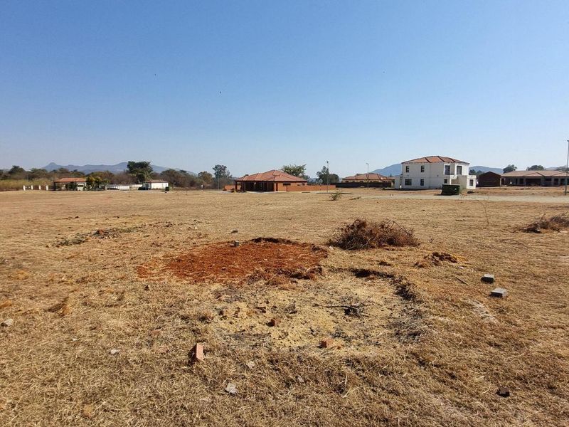 A vacant residential plot featuring dry grass and a brick boundary wall, situated in a developed neighborhood with surrounding houses.