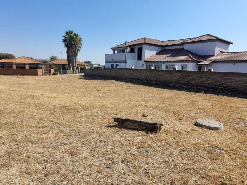 A view of a vacant residential plot featuring dry grass, a boundary wall, and a manhole cover, with neighboring properties visible in the background.
