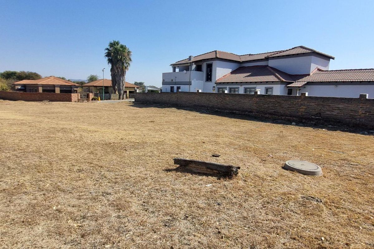 A view of a vacant residential plot featuring dry grass, a boundary wall, and a manhole cover, with neighboring properties visible in the background.
