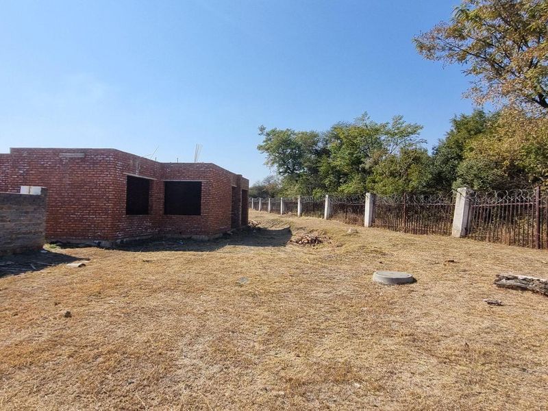 A view of a residential plot featuring an unfinished brick structure, perimeter walling with razor wire, and open land.