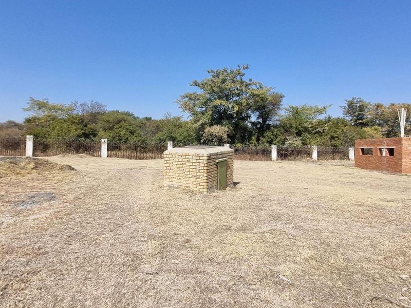 Vacant residential land featuring a partially constructed brick structure, a small utility enclosure, and perimeter fencing with mature trees in the background.