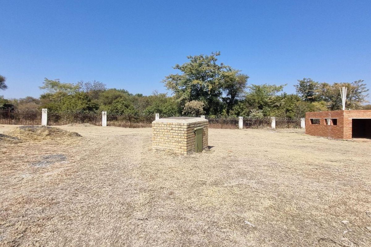 Vacant residential land featuring a partially constructed brick structure, a small utility enclosure, and perimeter fencing with mature trees in the background.