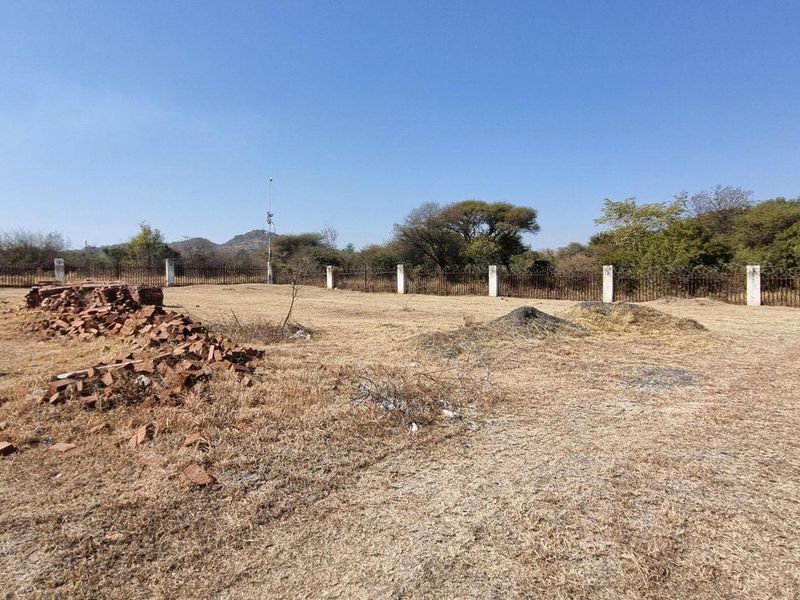 A vacant residential plot featuring perimeter fencing with concrete posts and a brick pillar, currently covered in dry grass and scattered construction debris.