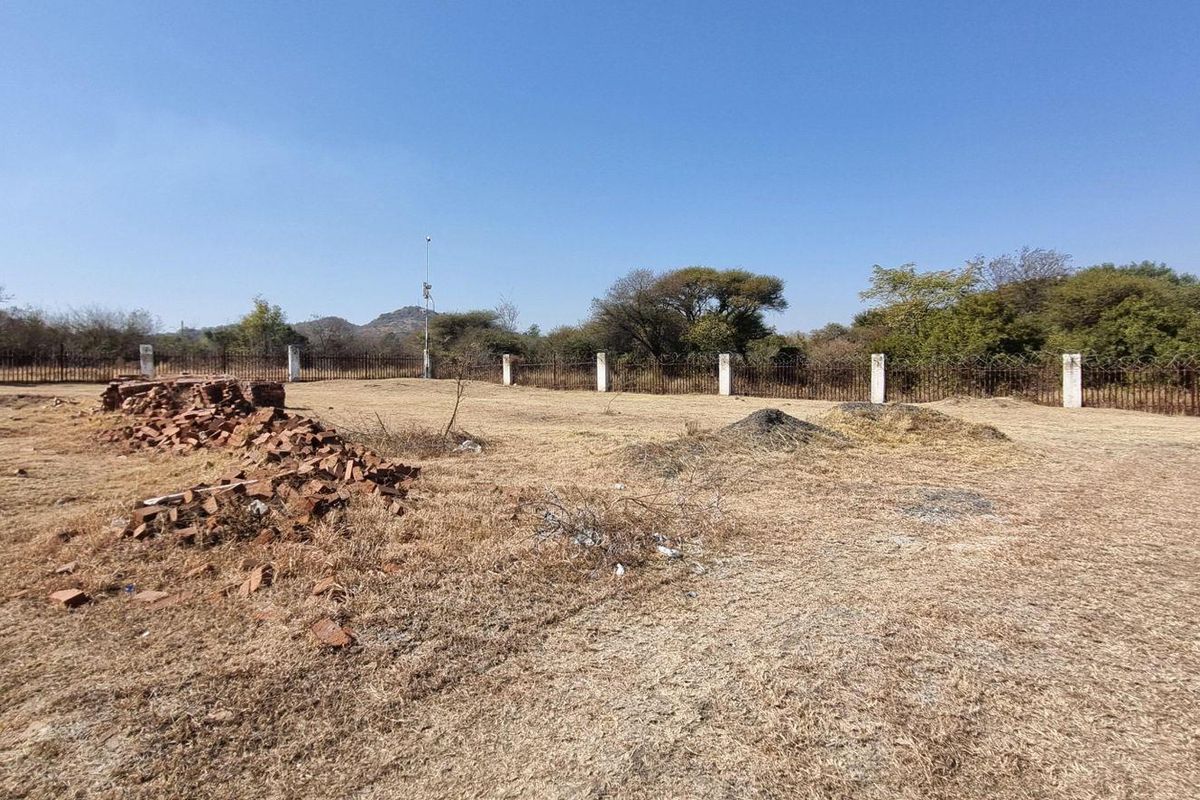 A vacant residential plot featuring perimeter fencing with concrete posts and a brick pillar, currently covered in dry grass and scattered construction debris.