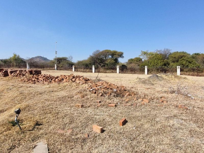 A vacant residential plot featuring perimeter fencing and concrete posts, with some scattered debris present on the dry grass surface.