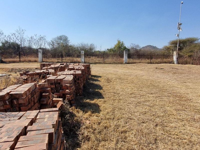 A vacant residential plot featuring dry grass, perimeter metal fencing with concrete posts, and a security camera pole.