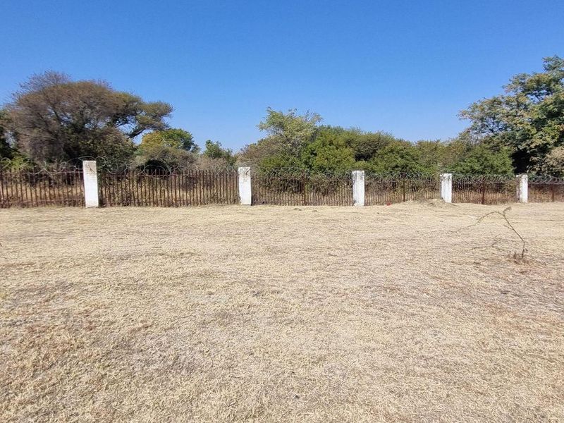A view of a vacant residential plot featuring dry grass and a perimeter fence with concrete pillars and security razor wire.