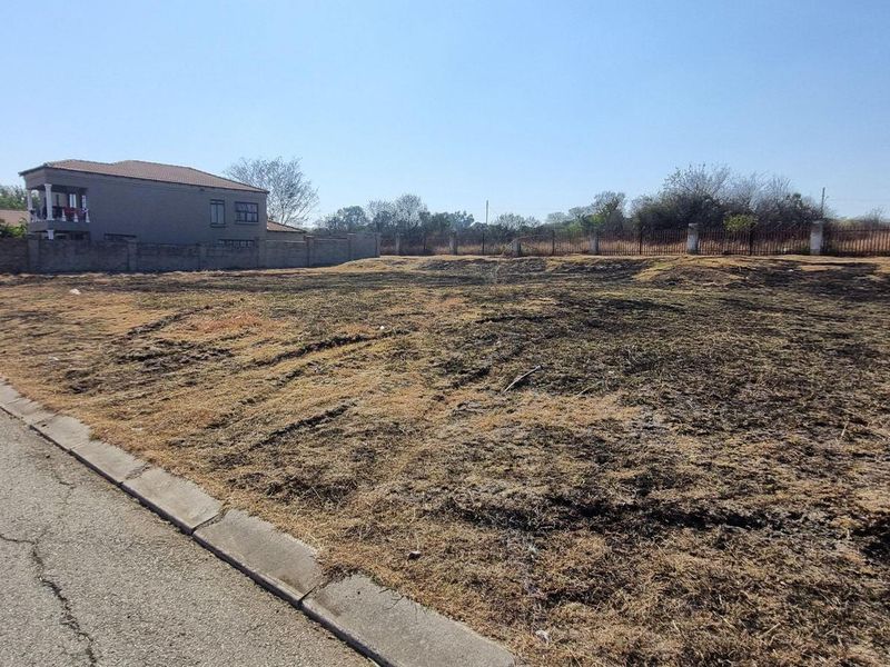 A vacant residential plot of land featuring a perimeter wall and metal fencing, situated along a paved street.