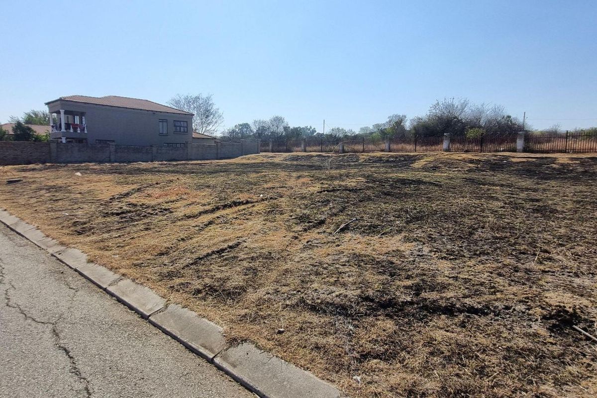A vacant residential plot of land featuring a perimeter wall and metal fencing, situated along a paved street.