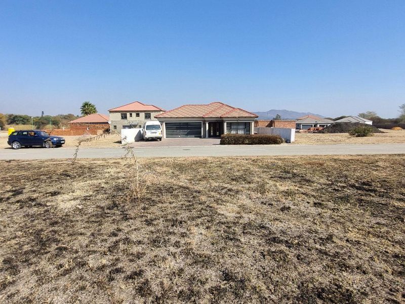 A wide-angle view of a vacant residential plot featuring dry, scorched grass with a paved road and neighboring houses in the background.